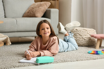 Cute little happy girl with pencil case, notebook and marker drawing while lying on floor at home