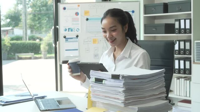 A young Asian businesswoman works alone at a desk with a laptop, stack of papers, tablet, and mobile phone. She looks happy and inspired, managing appointments and analyzing equity financing.