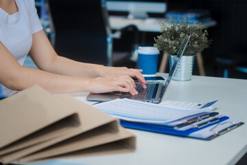 A young Asian woman works at her desk in an office room during the day. Surrounded by financial charts, files, and devices, she consults on credit reports, calculations, and credit risk assessments.
