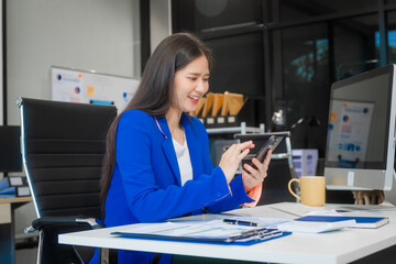 A young Asian woman works at her desk in an office room, managing time with a laptop, papers, and financial charts. She focused on consulting in various fields, from strategy to HR and legal.