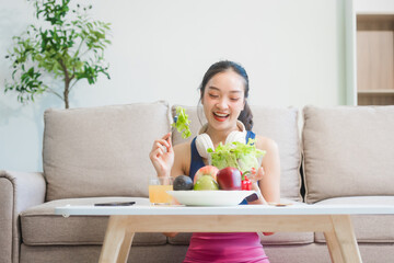 A young Asian housewife enjoys a healthy lifestyle at home, balancing fitness routines with relaxation. She sits on a modern sofa, eating a fresh salad and fruit while embracing healthy habits.