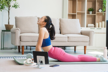 A young Asian housewife sits on sofa in her living room, using fitness app on her mobile with headphones. dumbbells and whey protein, she balances yoga, workouts, vegetarian diet and weight loss.