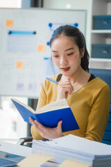 A young Asian businesswoman works alone at a desk with a laptop, stack of papers, tablet, and mobile phone. She looks happy and inspired, managing appointments and analyzing equity financing.