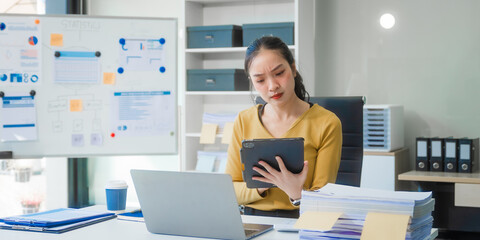 A young Asian businesswoman works alone at a desk with a laptop, stack of papers, and documents. She looks serious and stressed, analyzing financial losses and equity management under daylight.