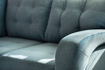Soft blue velour sofa with quilted cushions and stitched armrest in shadows and sunlight. Part of...
