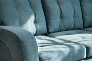 Soft blue velour sofa with quilted cushions and stitched armrest in shadows and sunlight. Part of the sofa with a soft back and seat close-up with copy space.