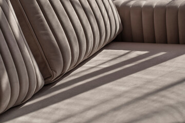 Soft beige velour sofa quilted with straight lines of stitching in shadows and sunlight. Part of the sofa with armrest and pillows close-up with copy space.