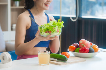 A young Asian housewife enjoys a healthy lifestyle at home, balancing fitness routines with relaxation. She sits on a modern sofa, eating a fresh salad and fruit while embracing healthy habits.