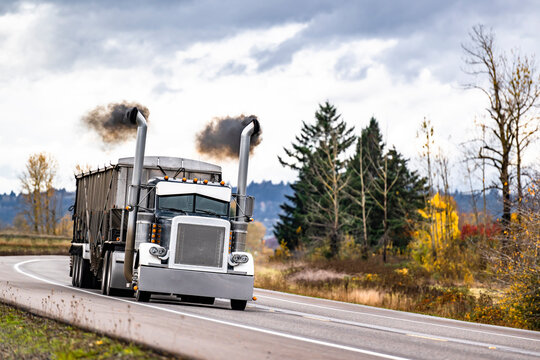 Black big rig classic semi truck with with thick black smoke coming from the exhaust pipes transporting cargo in bulk semi trailer running on the road polluting the environment