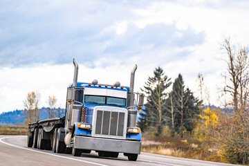 Blue classic American idol big rig semi truck tractor with empty flat bed semi trailer running on the turning autumn road