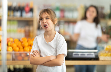 Angry child is standing in grocery store, capricious and sulky silence. Girl makes faces and ignores conversation with her mother, parent screams and is indignant in background
