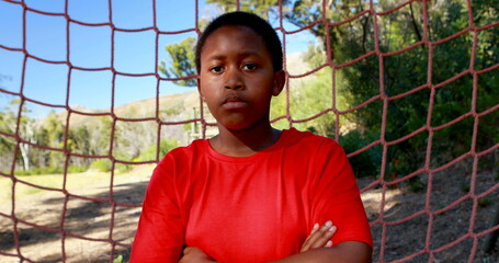 Determined boy standing with arms crossed against net during obstacle course in boot camp