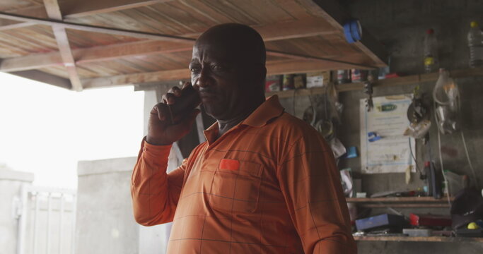 Front view of an African male owner of a panel beater workshop in a township, talking on the smartph