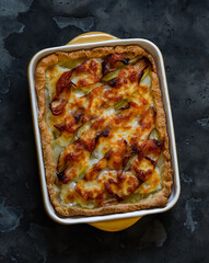 Pie with potatoes, bacon and mozzarella cheese in a baking dish on a dark background, top view