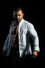 Sexy male model with hot naked body in white shirt. A sexy man in an unbuttoned white shirt on black studio. Handsome muscular man with naked torso and shirt. Sexy Guy with with open shirt.