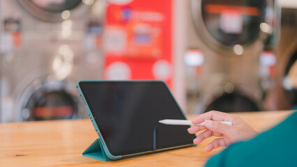 Tablet on wooden table in laundromat, hand using stylus, blending productivity with everyday chores...
