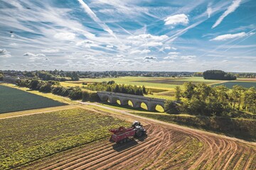 Fototapeta premium Vibrant agricultural landscape under a sprawling sky with a tractor in action near an old stone bridge