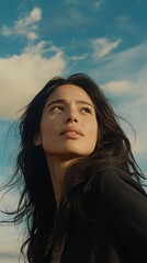 Fototapeta premium A woman with dark hair looks upwards against a backdrop of a clear, vibrant sky, exuding serenity and introspection.