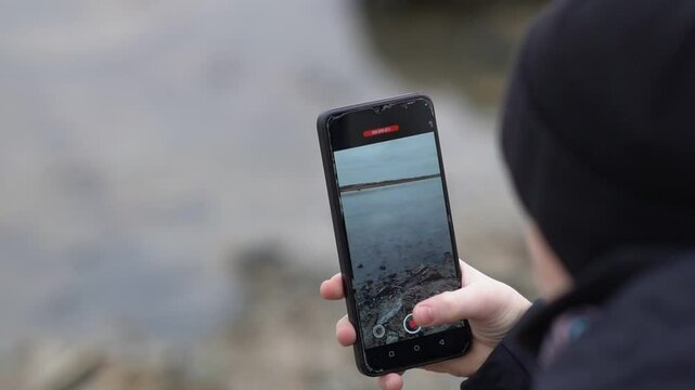Cropped close-up shot of a boy with a broken arm, wrapped in a white plaster cast. A teenager uses a phone in a park near a pond
