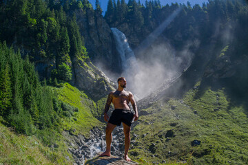 Obraz premium Man tourist on summer vacation. Full length photo of Hispanic man with naked sexy torso on summer vacation. Hiker guy outside. Tourist man walking on the waterfall landscape. Man Traveling and nature.