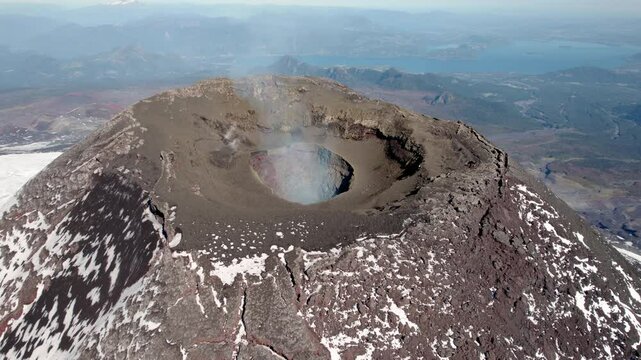 A breathtaking view from the summit of a volcano reveals a large crater surrounded by rocky terrain, with a serene lake visible in the distance against a clear blue sky.