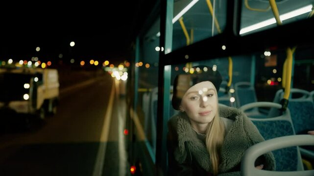 A beautiful young woman looks out the window of a bus going through the night city. 