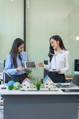 Two businesswomen sit at a table in the office, discussing a model village with windmills,green trees,sustainable living,renewable energy, circular economy,eco-friendly cities for the world's future