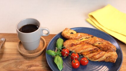 Crisp twist bread on blue plate with fresh basil and ripe cherry tomatoes, accompanied by hot black coffee on rustic wooden board, creating cozy breakfast ambiance.