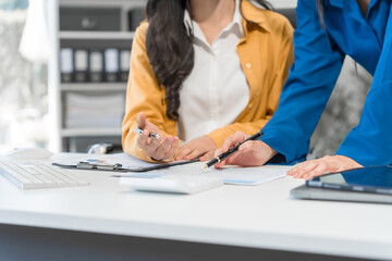 Two Asian businesswomen work on a laptop in an office, pointing at the screen, talking, and smiling with joy as they celebrate positive results of their business success.