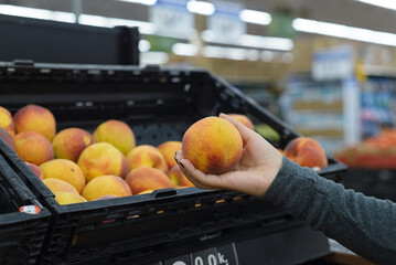 Female hand holding a peach next to a shelf in a supermarket.