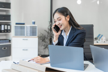 Two Asian businesswomen work on a laptop in an office, pointing at the screen, talking, and smiling with joy as they celebrate positive results of their business success.