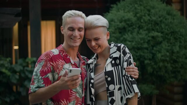 A young blond man with tattoos and a beautiful blond woman with tattoos hold a phone outdoors. They win a prize in a mobile app. Overjoyed, they can't contain their emotions. He hugs and kisses her.