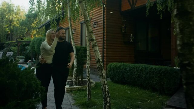 A couple walks by pool in country house garden, hugging and discussing. They pass the camera at the end. She wears a white top, jeans, and black high heels; he wears black pants and a black T-shirt.
