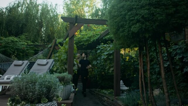 A man holds his girlfriend's hand while touring his country house. They walk by the pool, and she steps onto a ledge in black high heels. He supports her as she walks along it. At the end they kiss.