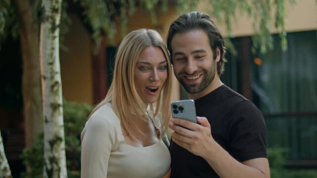 The couple holds a phone, shocked by their mobile app win. Truly overjoyed, they show the smartphone green screen toward the camera, celebrating their truly unexpected prize on the phone.