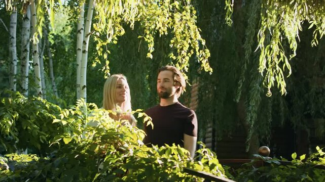 Two young people&mdash;a blonde woman and a brunette man&mdash;walk through a park. They navigate dense bushes and emerge onto a bridge. The couple gently embraces and gazes into the distance.