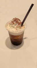 close up of a cold coffee drink in a clear plastic cup, this drink is topped with whipped cream, sprinkled with cocoa powder, and a black straw is inserted into it isolated on wood background