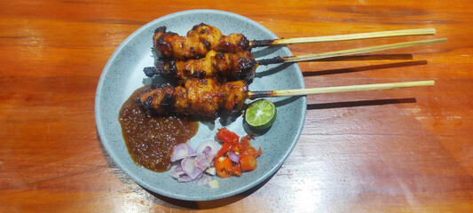 a plate of grilled chicken satay merah  culinary, served with additional honey sauce, sliced ​​shallots, chopped red chilies, and halved lime, on a gray plate, isolated on wood background