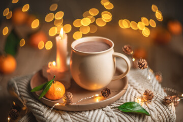 Christmas hot chocolate with tangerines and burning candles on a stack of knitted sweaters creates a cozy winter atmosphere with warm bokeh lights