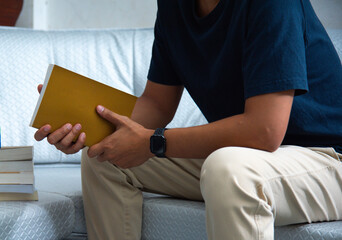 A man is seated comfortably on a sofa, engaged in reading a book. His attire is casual yet neat, such as a shirt and trousers, suggesting a moment of leisure within a serene environment.