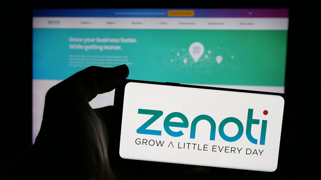 Stuttgart, Germany - 11-15-2024: Person holding mobile phone with logo of US salon software company Soham Inc. (Zenoti) on screen in front of web page. Focus on phone display.