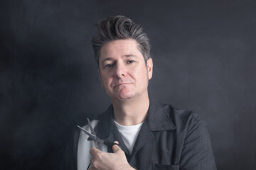 Portrait of a mature man with toupee hairstyle, looking at camera serene and serious expression, holding a pair of scissors on black and smoke background. Male hairdressing concept