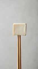 A single piece of tofu held by a pair of chopsticks, centered on a light grey background