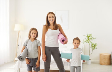 Fototapeta premium Happy woman in sport clothes standing in bedroom with her children, preparing to home workout together. Cheerful sporty mother and kids holding yoga mats and looking at camera. Healthy family concept.
