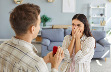 Young man with a red gift box proposing to his happy excited girlfriend standing in living room at home. Male person giving surprise gift with engagement ring or jewelry to pretty girlfriend.