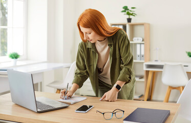 Portrait of a young redhead business woman in casual clothes working with laptop computer in office...