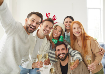 Portrait of young happy friends group standing in living room at home celebrating Christmas holiday or New Year and looking cheerful at camera making selfie with glasses of champagne in hands.