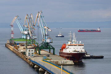 Naklejka premium Sea trade port in the town of Korsakov. Sakhalin Region, Far East of Russia. View of the seaport, a moored passenger ship, port cranes on the pier, cargo ships and tugs. Aniva Bay, Sea of ​​Okhotsk.