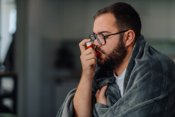 Sick man using inhaler at home wrapped in blanket