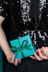 Woman in black sequin dress holding blue gift box behind her back on green velvet curtain...